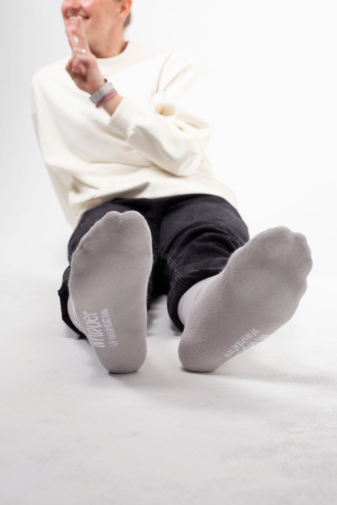 Cremefarbene Socken von Whisper of Inspiration mit Logo auf der Sohle an einer sitzenden Person vor hellem Studiohintergrund.