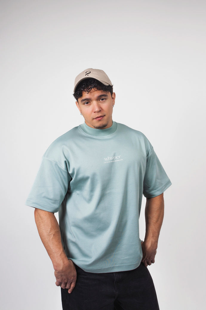 Model trägt das Whisper of Inspiration T-Shirt in Sage Green mit oversized Passform und beiger Cap vor hellem Studiohintergrund.